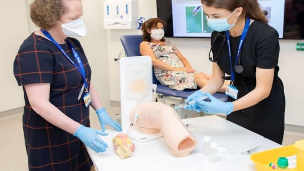 Dr Bernadette Lynch, Consultant Rheumatologist at University Hospital Galway, guiding a Medical Trainee through a simulated knee aspiration. Patient on stretcher in background. All wearing medical masks.